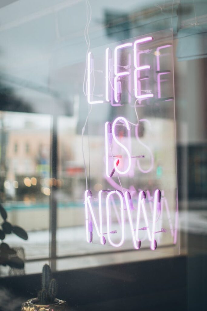 pexels photo 3811074 Urban scene with a glowing 'Life is Now' neon sign reflecting in a window, conveying a modern, motivational vibe.